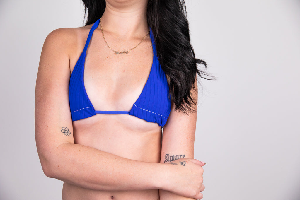 Person wearing a blue bikini top with tattoos on arms against a plain background