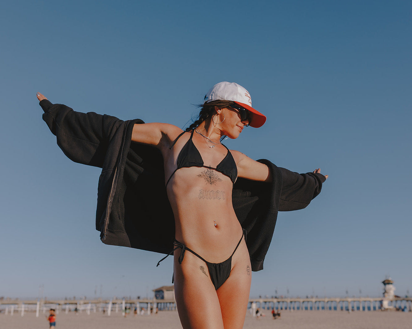 Woman in a black bikini and cap standing on a beach with a clear blue sky.